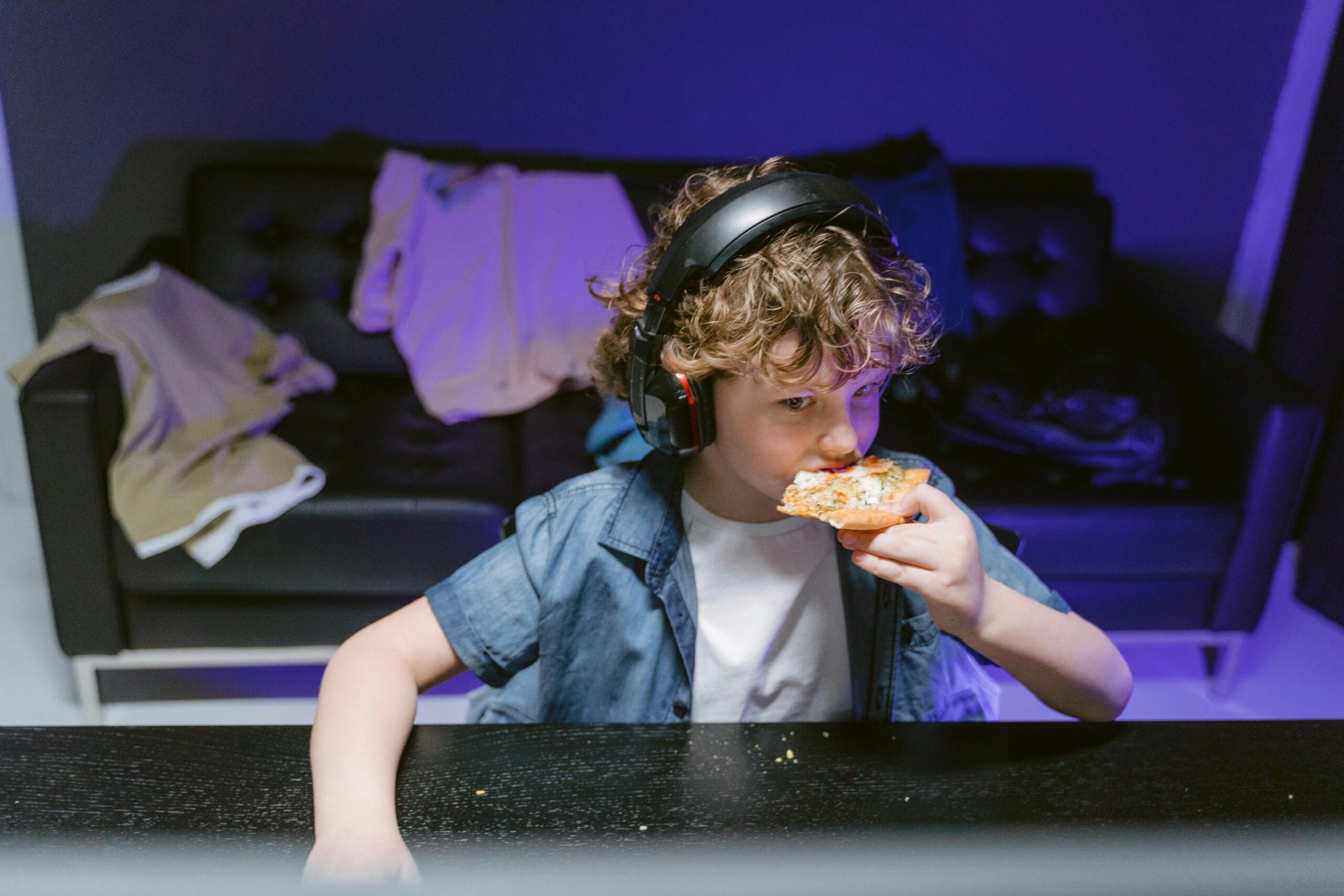 Curly-haired boy enjoying a slice of pizza while wearing headphones indoors.