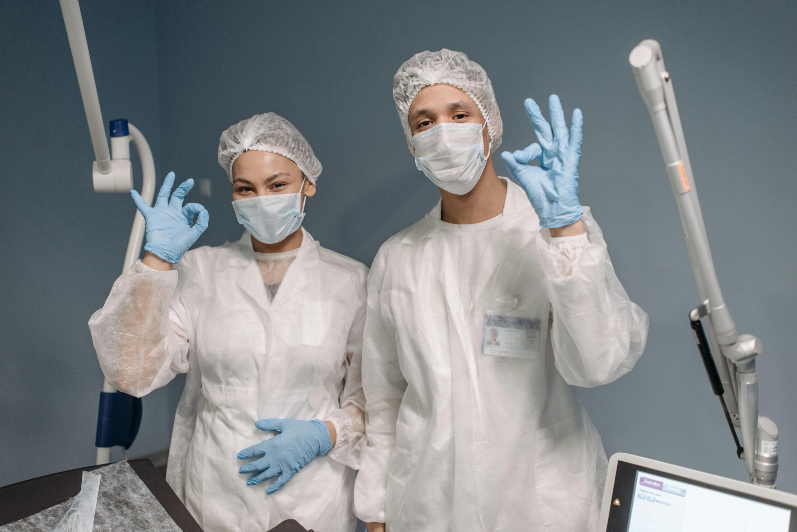 Two healthcare workers in PPE with masks and gloves giving an OK sign in a medical setting.