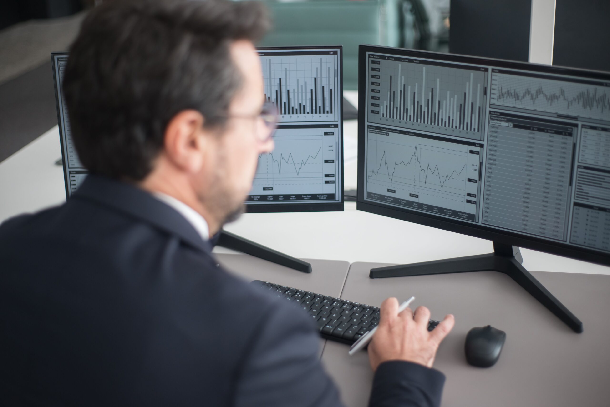 A businessman in a suit analyzing financial charts and graphs on two computer monitors indoors.